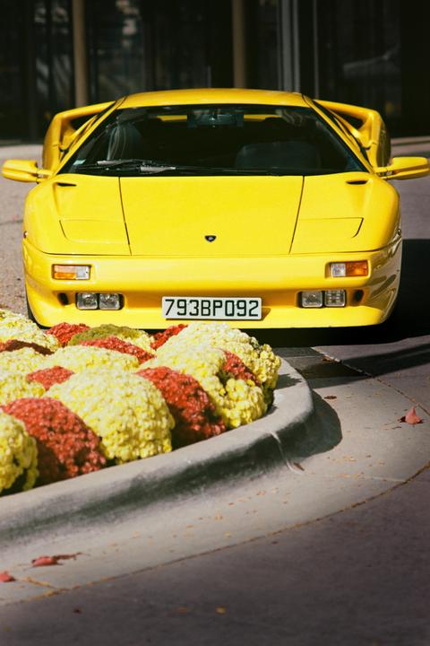 A bright yellow sports car is parked behind a planter filled with red and yellow flowers.