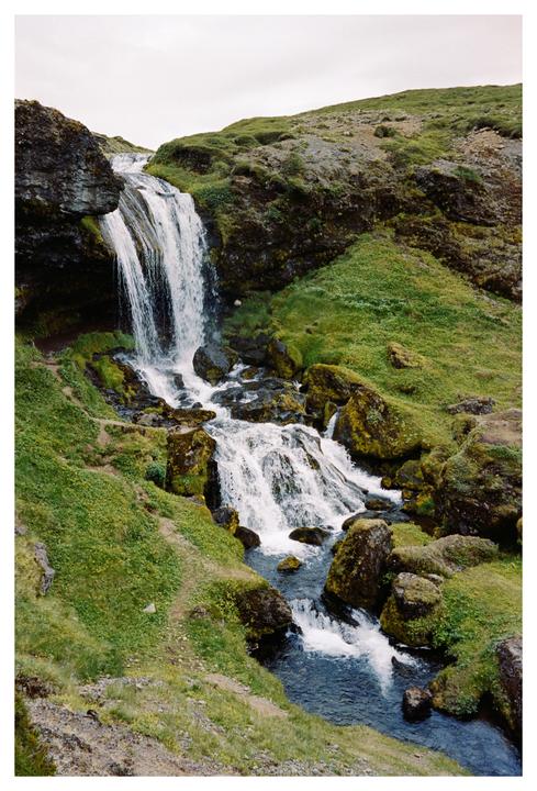 A waterfall cascades down a mossy, rocky hillside with a sheep visible near the top of the falls.