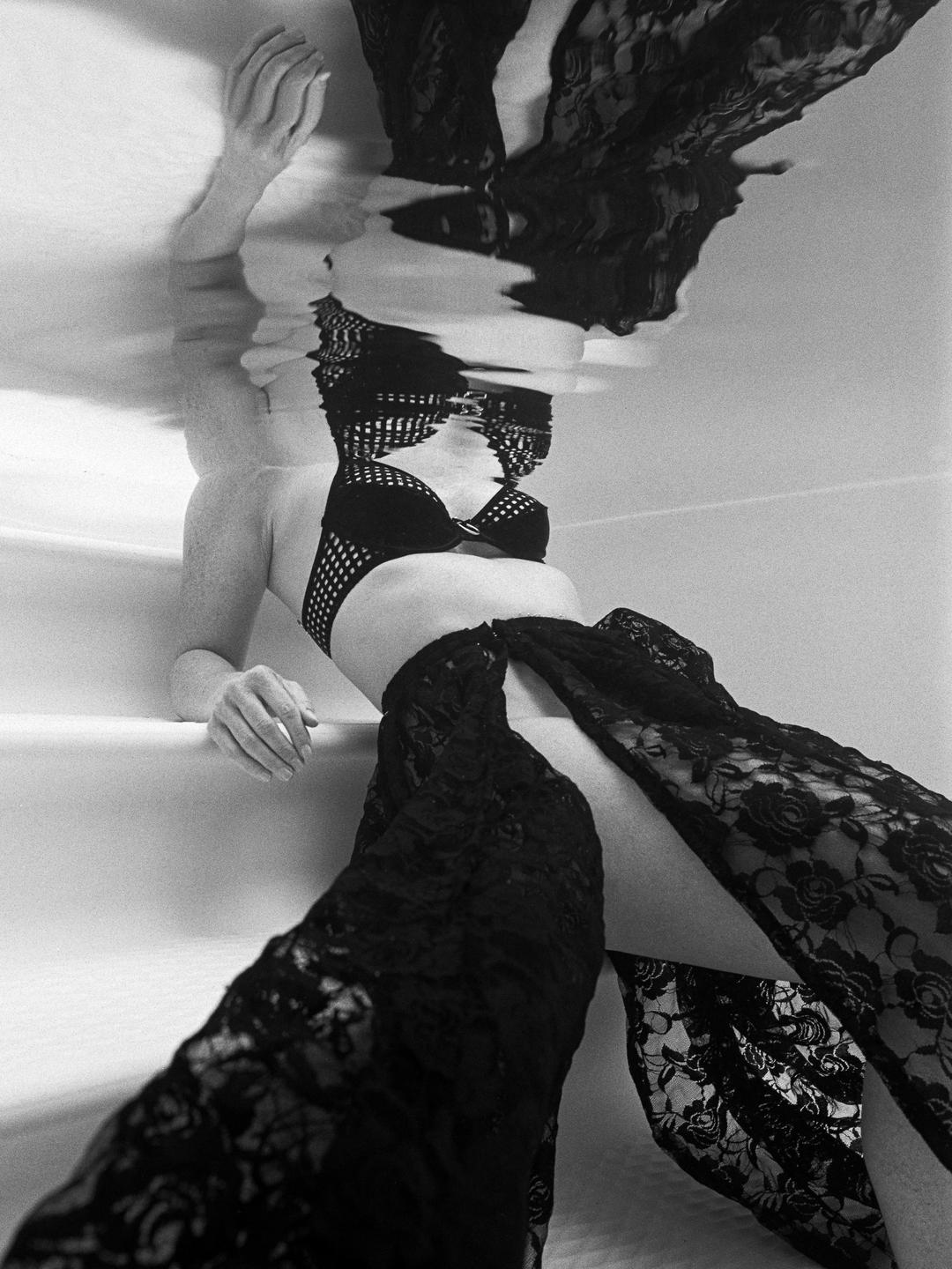 A woman in a black lace outfit is partially submerged in water, with her reflection visible on the surface.