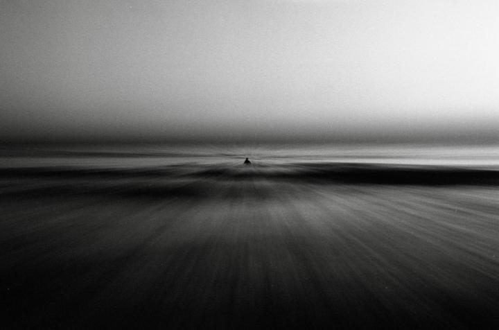 A lone surfer sits on a longboard in the ocean under a hazy sky.