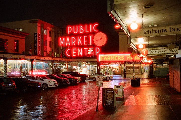 A street at night is illuminated by red neon signs and reflections on the wet pavement.