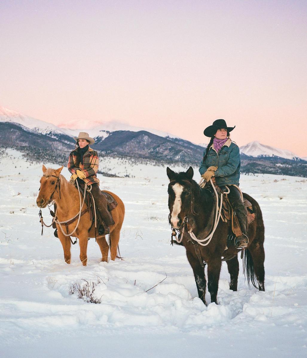 Two women on horseback stand in a snowy field with mountains in the background.