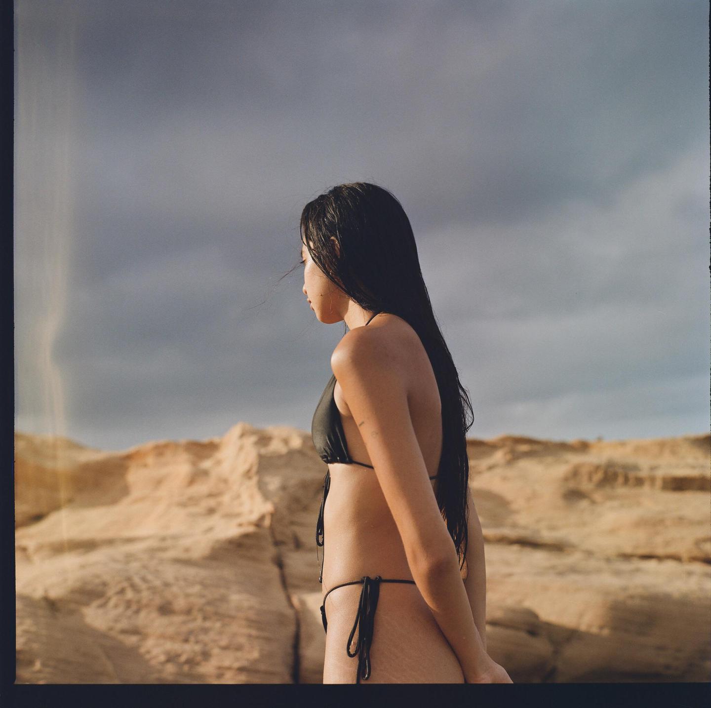 A woman in a bikini stands in a desert landscape with a cloudy sky.