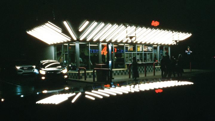 A brightly lit fast food restaurant with several people standing outside is visible at night.