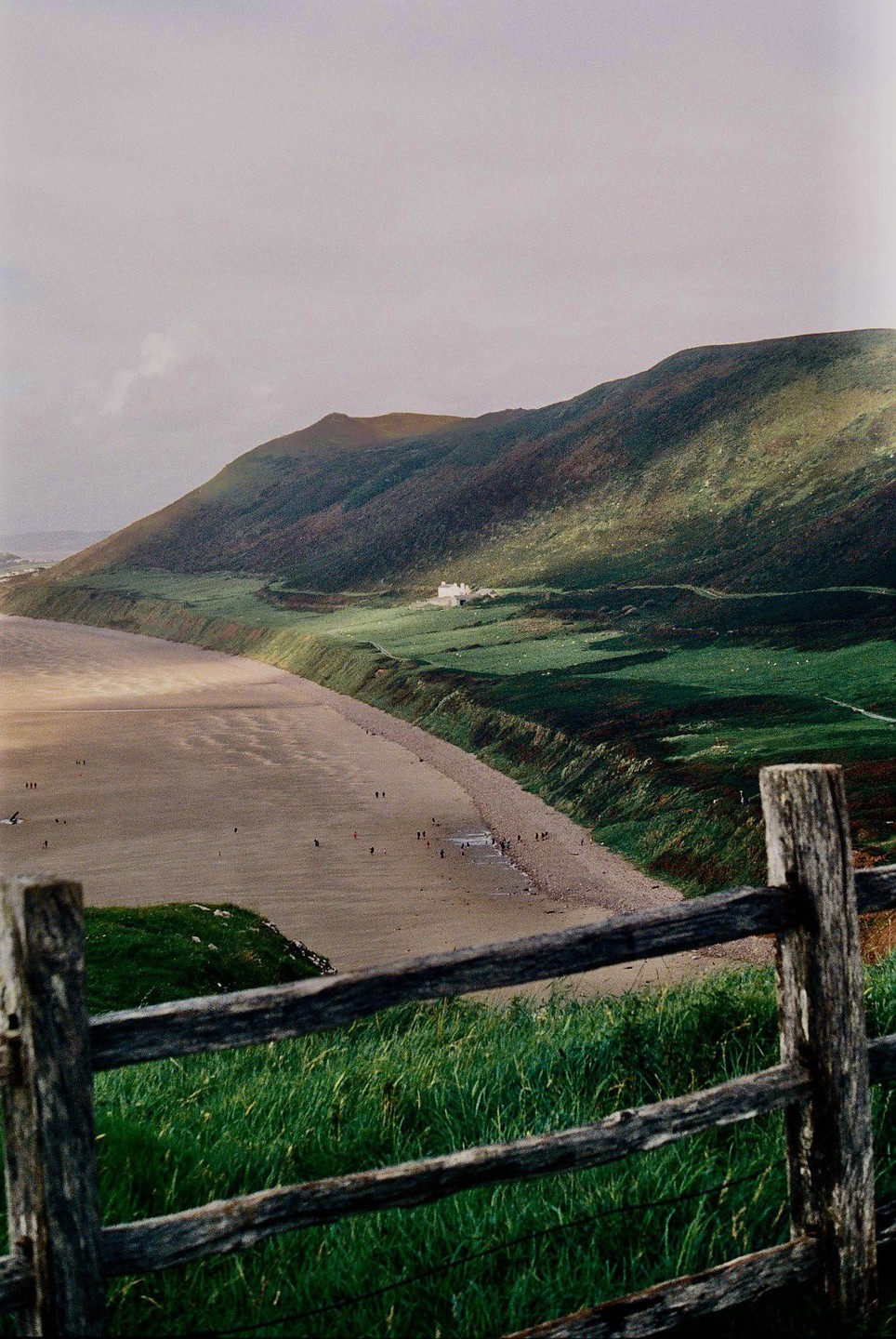 A wooden fence in the foreground overlooks a beach with people, a green hillside, and a house in the distance.