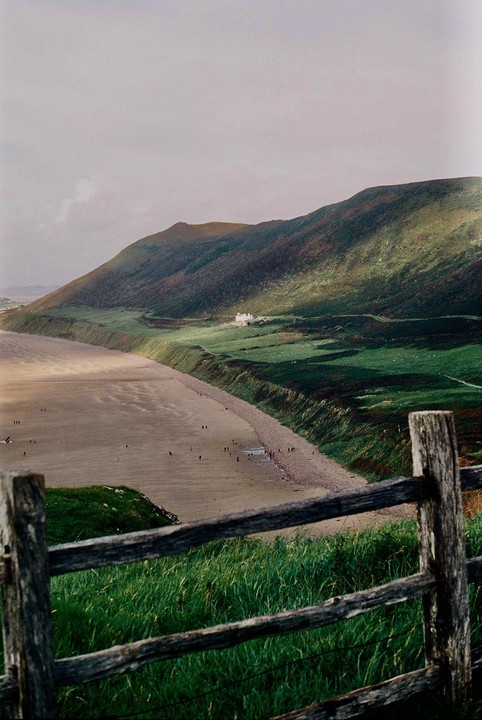 A wooden fence in the foreground overlooks a beach with people, a green hillside, and a house in the distance.