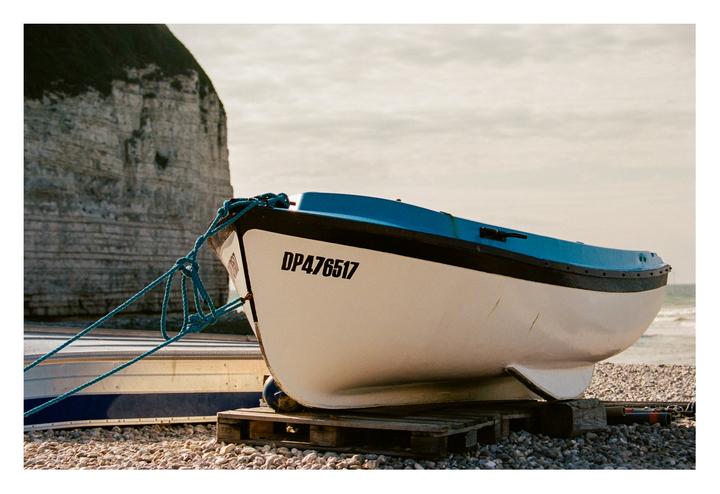 A white and blue boat sits on a rocky beach with a large cliff in the background.