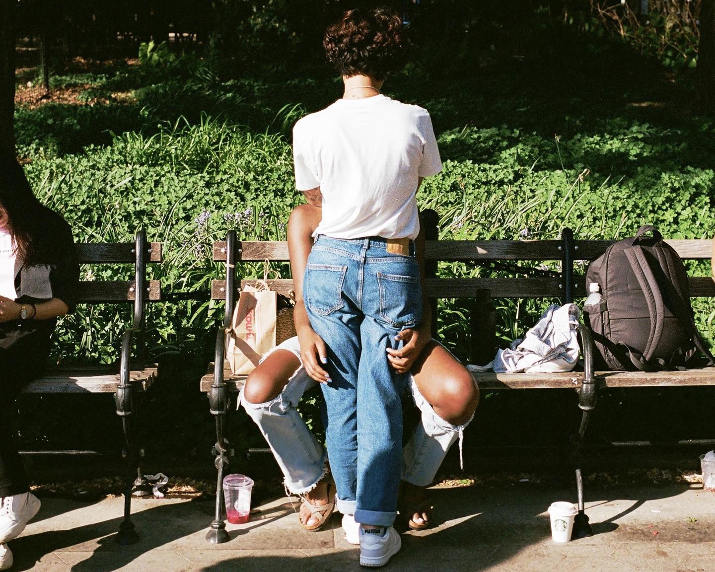 A couple sits on a park bench with a woman standing in front of a man, both wearing jeans.