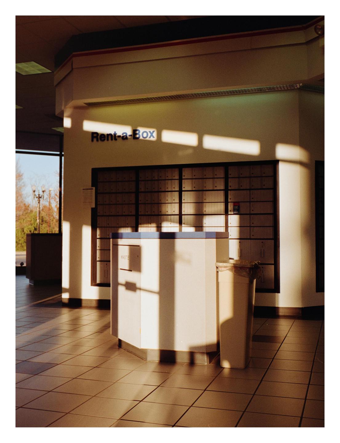 Sunlight streams into a post office lobby with rows of mailboxes and a counter.