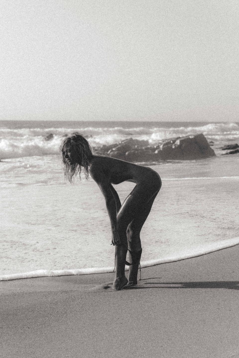 A woman bends over on a sandy beach with waves and rocks in the background.