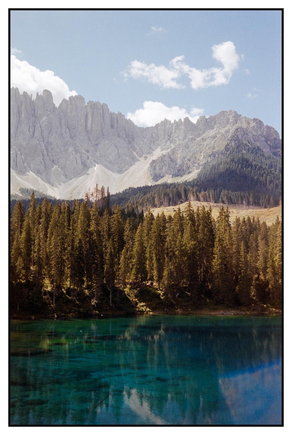 A turquoise lake reflects a forest and mountains under a blue sky with clouds.