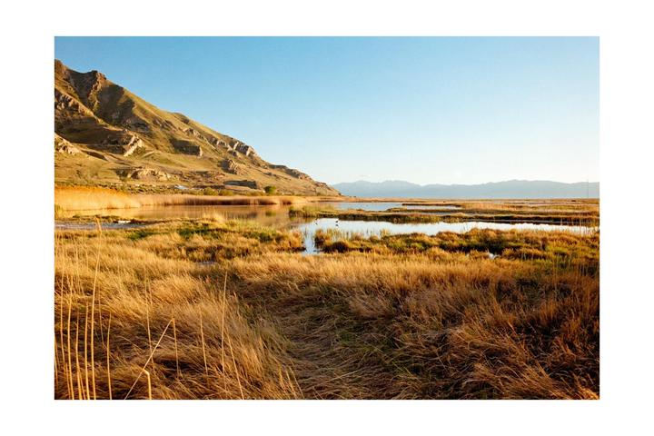 A marshy landscape with a large mountain on the left and smaller mountains in the distance under a clear sky.