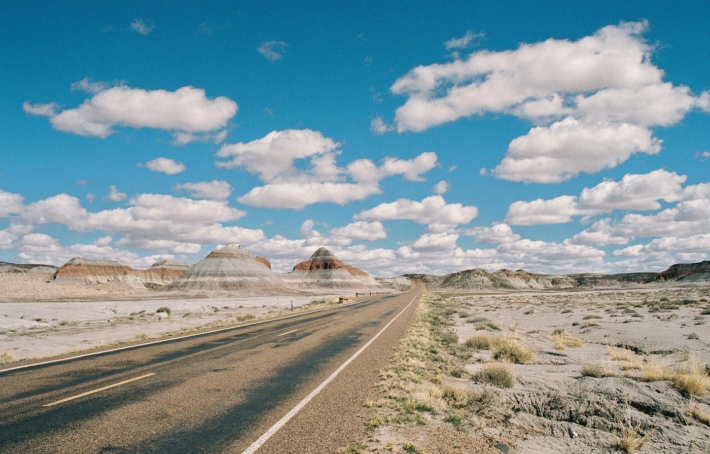 A long, empty road stretches through a desert landscape with colorful, layered hills under a bright blue sky with scattered clouds.