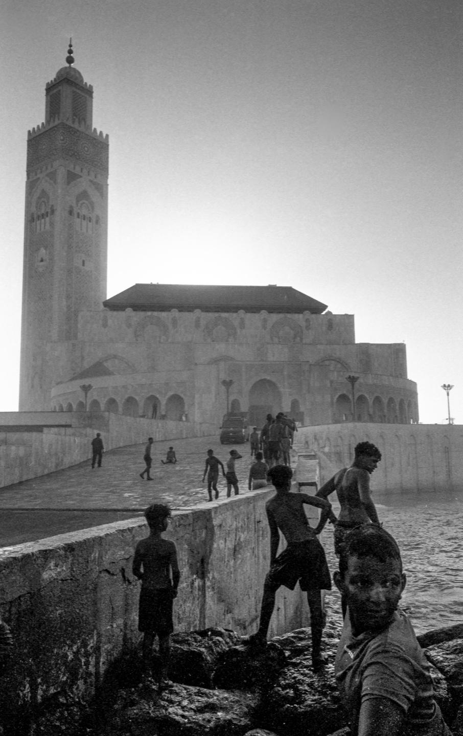 A group of boys stands on rocks by the water with a large mosque in the background.