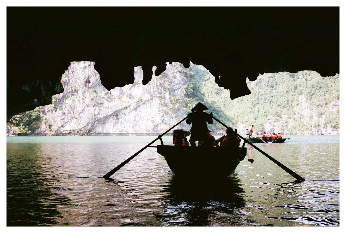 A person in a conical hat rows a boat with passengers out of a dark cave toward a body of water with limestone cliffs and other boats.
