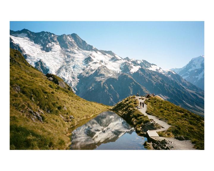 A glacial lake reflects snow-capped mountains under a clear sky, with a path leading to a group of people.