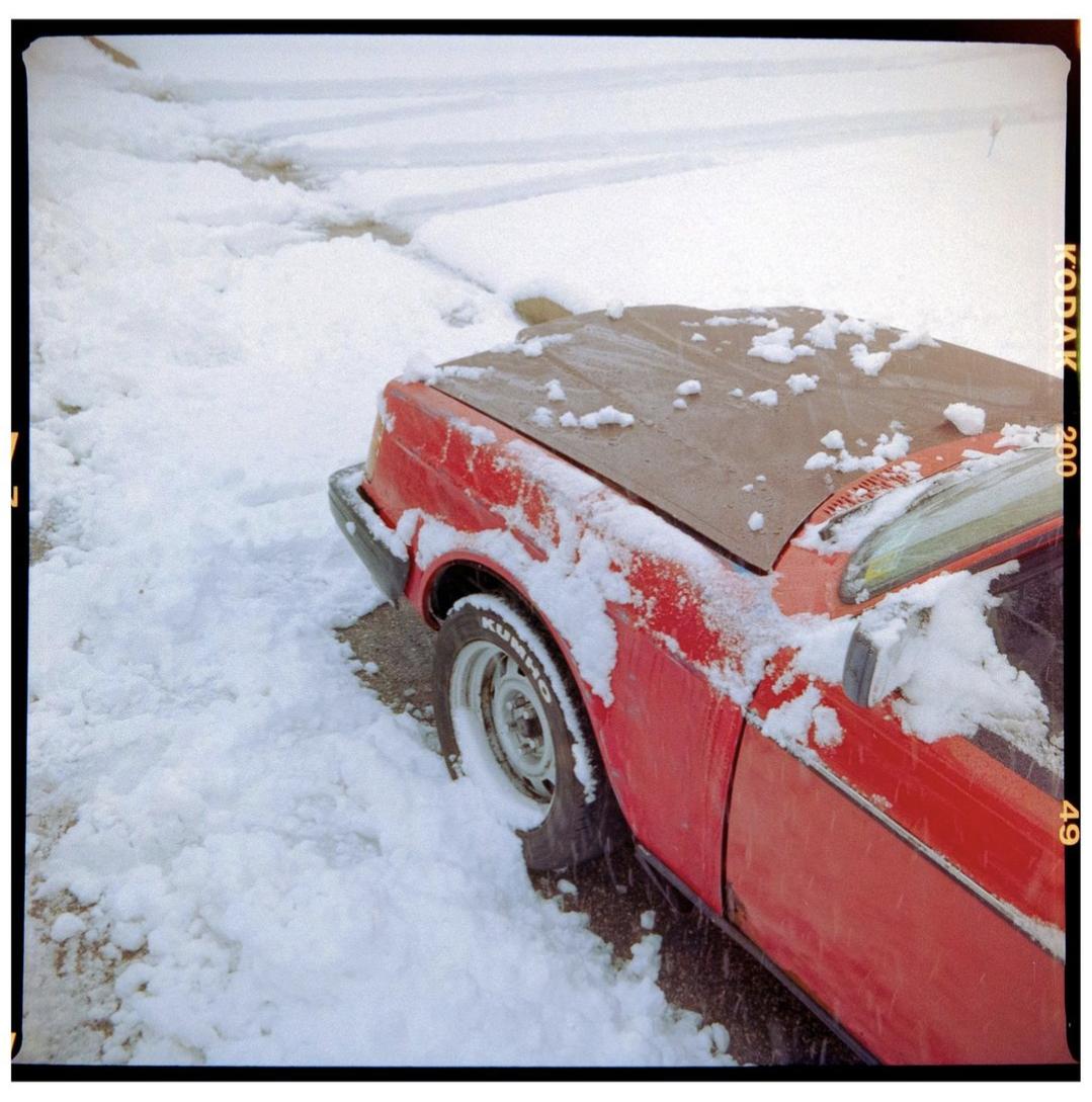 A red car with a dark hood is parked in a snowy area with tire tracks visible in the snow.