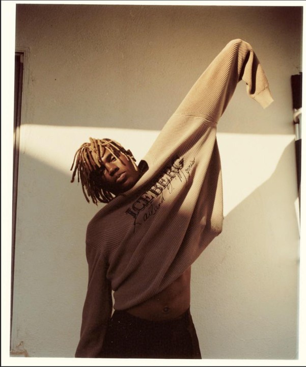 A man with dreadlocks poses with a sweater pulled over his head and one arm raised, casting a shadow on a white wall.