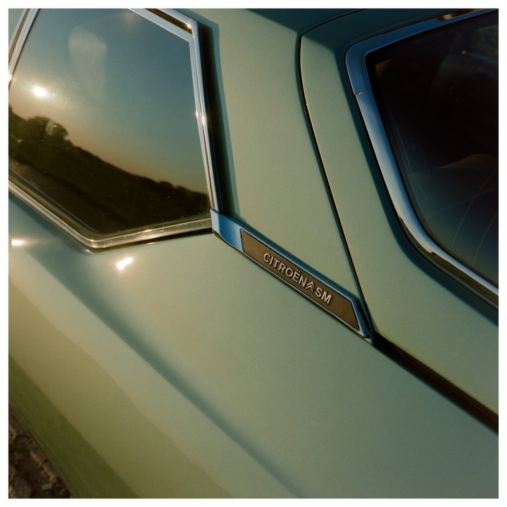 The side of a vintage Citroën SM car is visible with a reflection of the sun in the window.