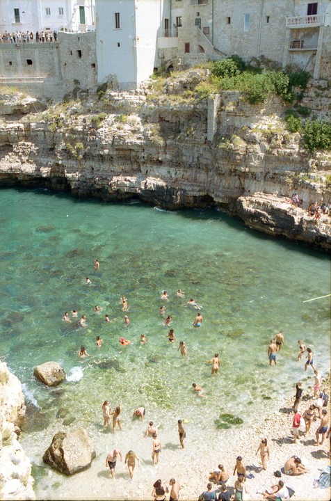 A crowd of people swim and relax on a small beach surrounded by cliffs and buildings.