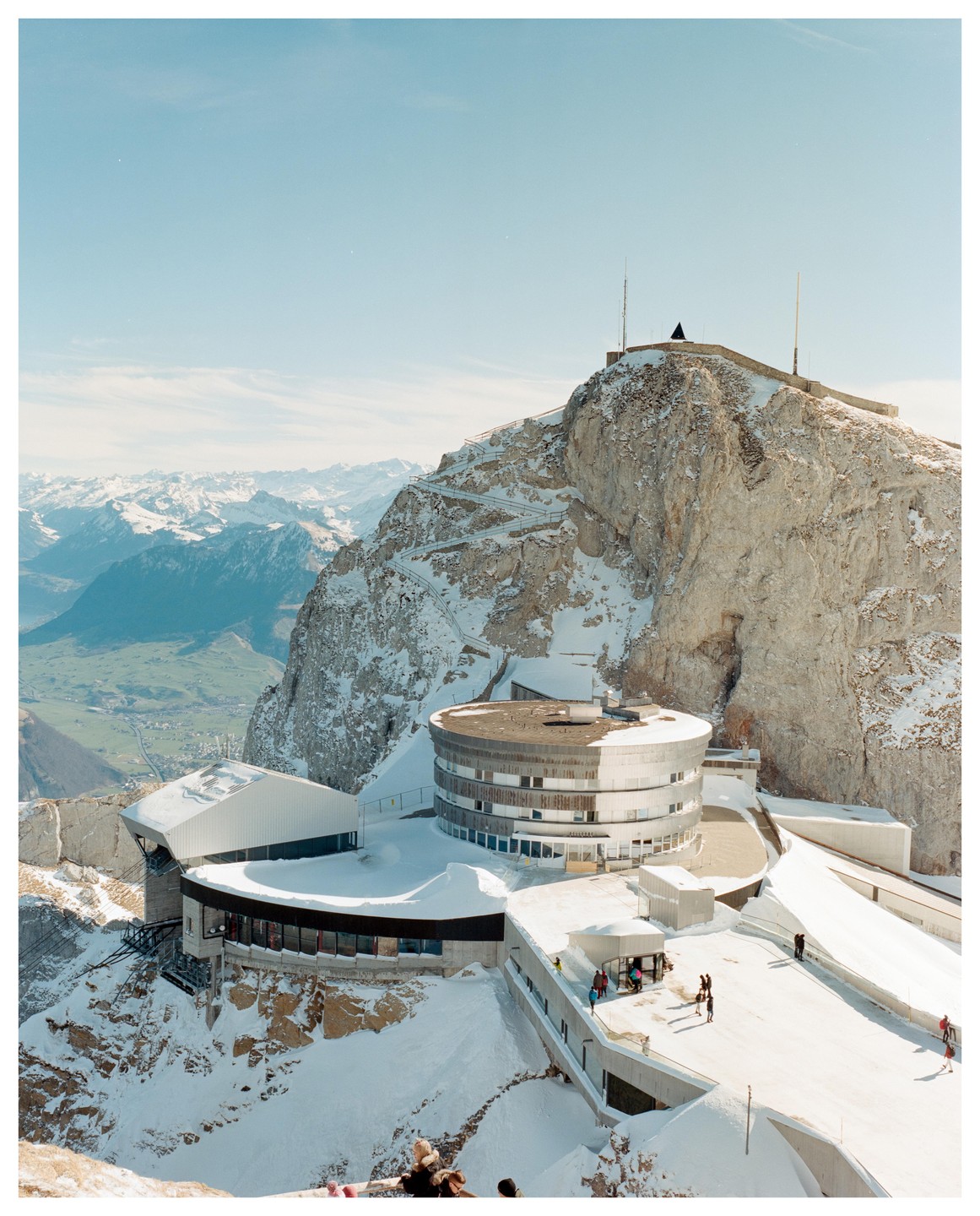 A modern building complex is nestled into a snow-covered mountain with a winding path leading to its peak.