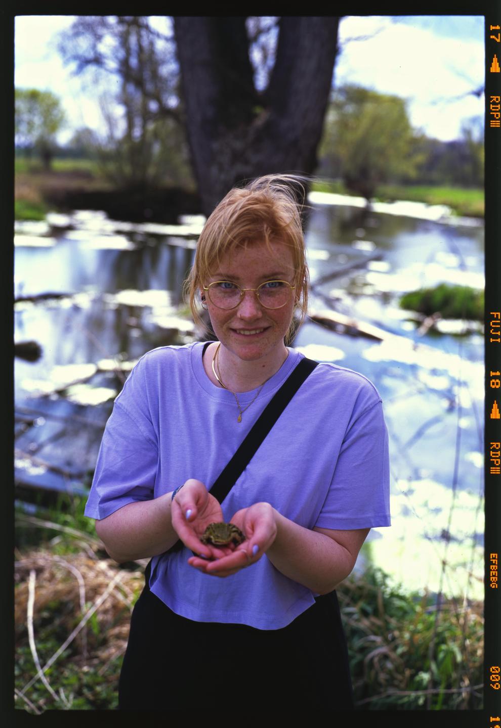 A woman with glasses holds a small frog in her hands while standing in front of a body of water.