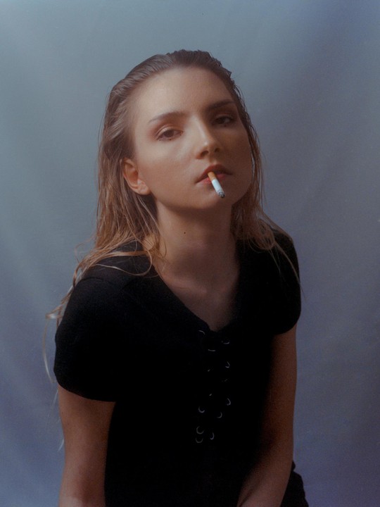 A woman with wet hair and a black shirt holds a cigarette in her mouth while looking at the viewer.