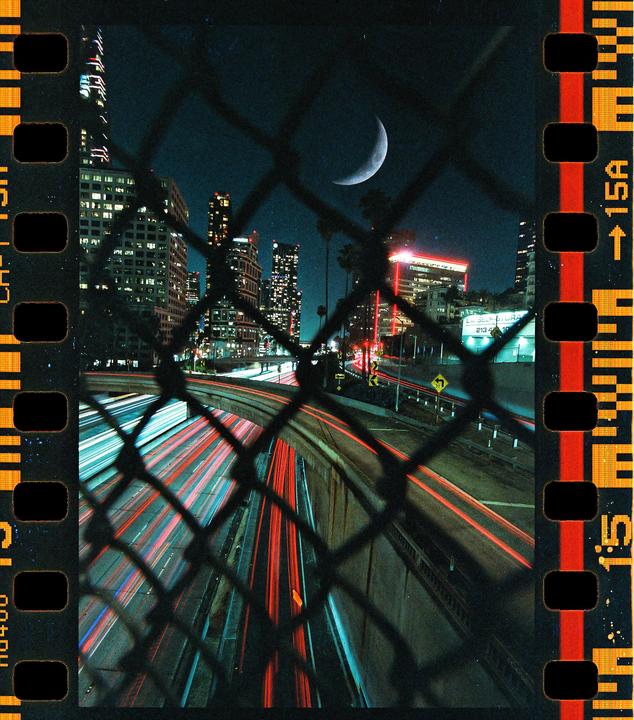A crescent moon hangs over a city skyline and a freeway with light trails, viewed through a chain link fence.