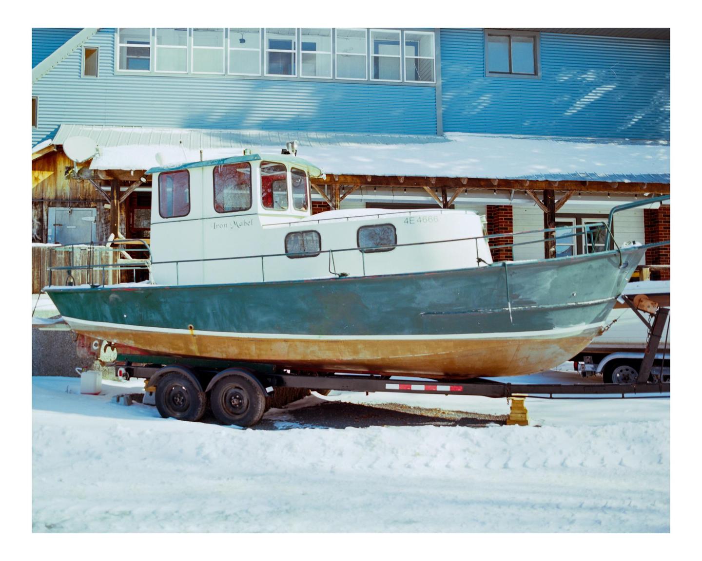 A boat named "Iron Mabel" sits on a trailer in the snow in front of a building.