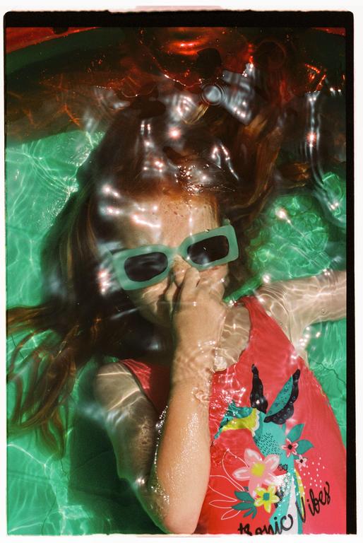 A girl with long red hair and green sunglasses floats in a swimming pool, covering her mouth with her hand.