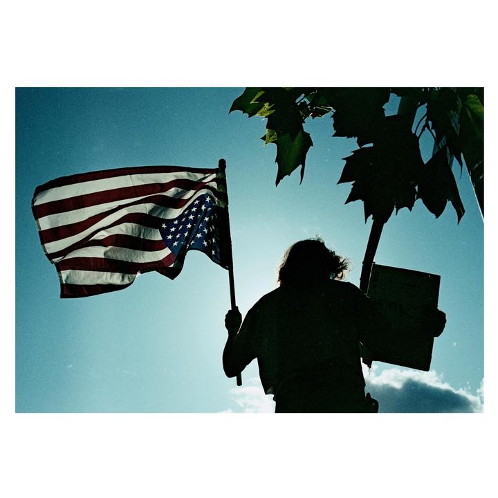 A person holds an American flag and a sign, silhouetted against a bright sky with some clouds and tree leaves.