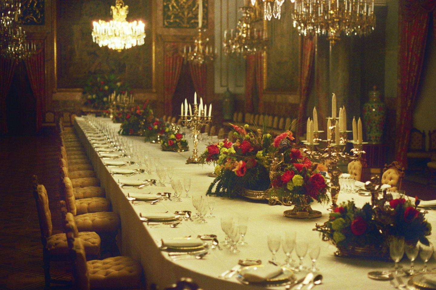 A long dining table is set with formal place settings, floral arrangements, and candelabras in a grand hall.