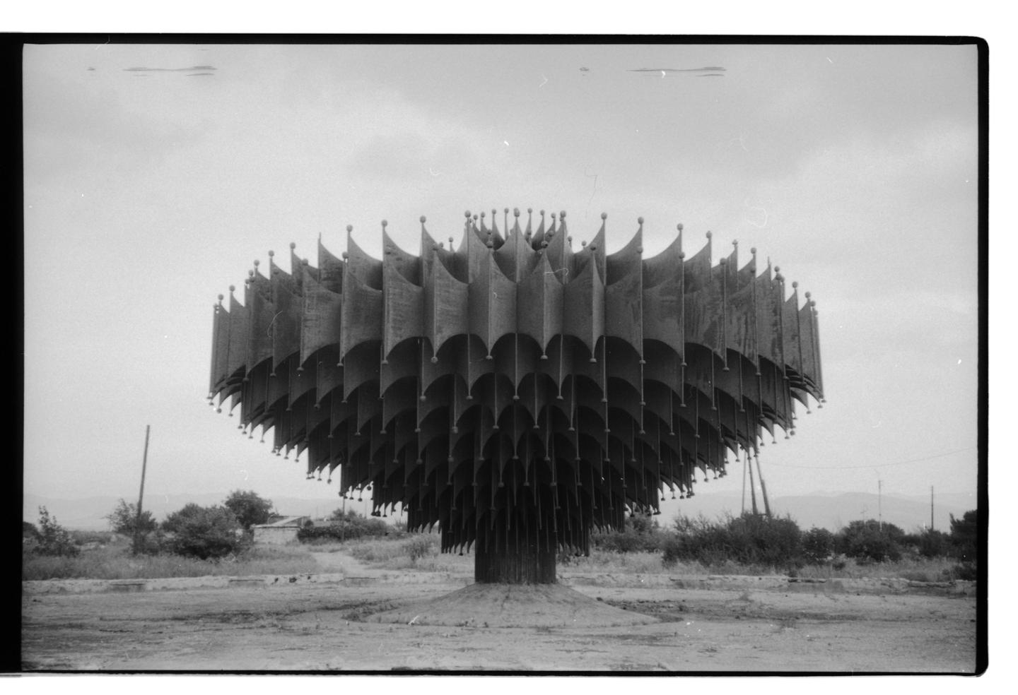 A large, round, metal sculpture with many flag-like elements stands in a barren landscape under a cloudy sky.