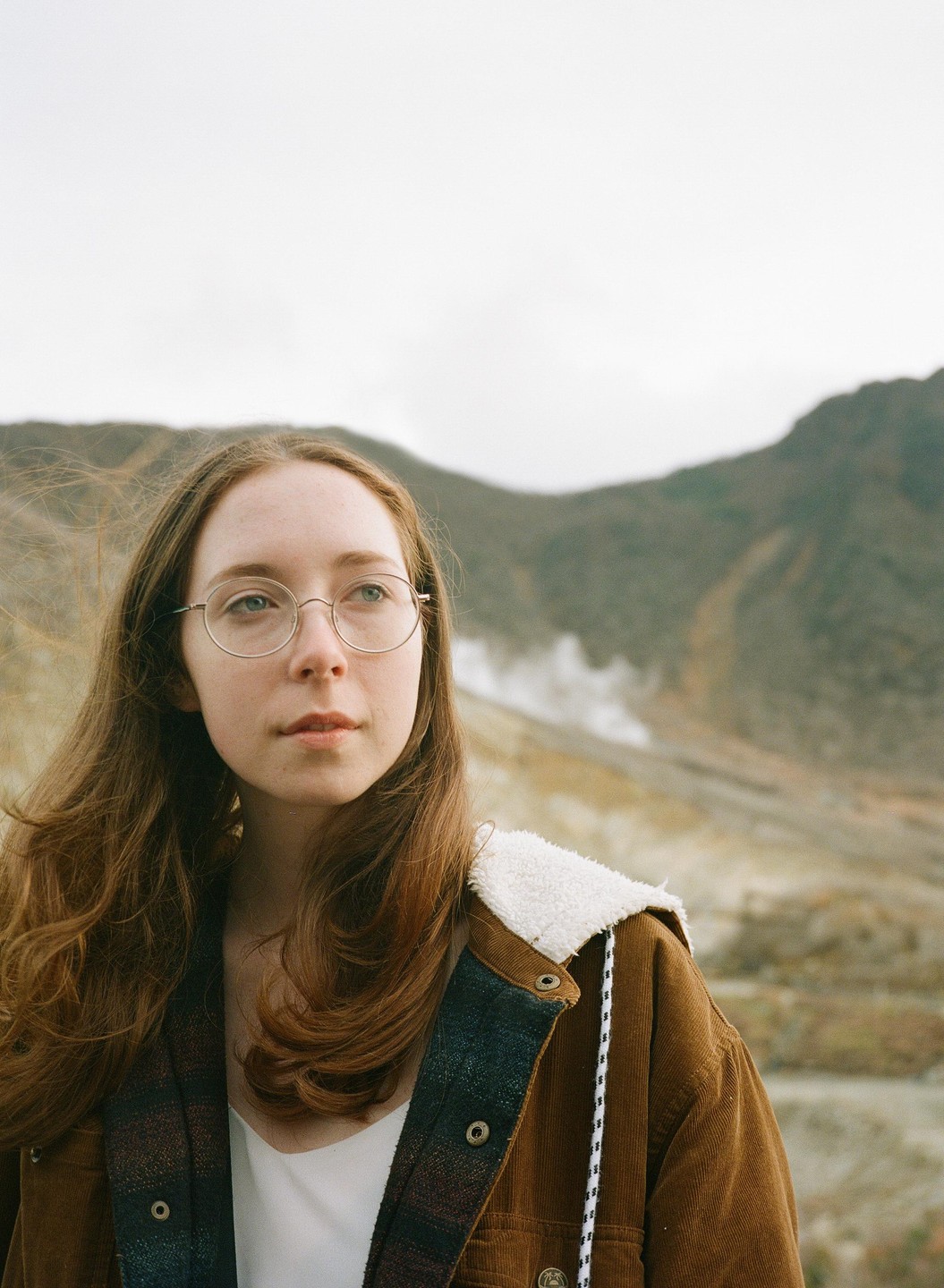 A woman with long hair and glasses wears a brown jacket with a white sherpa collar and looks to the right, with a mountain and steam in the background.