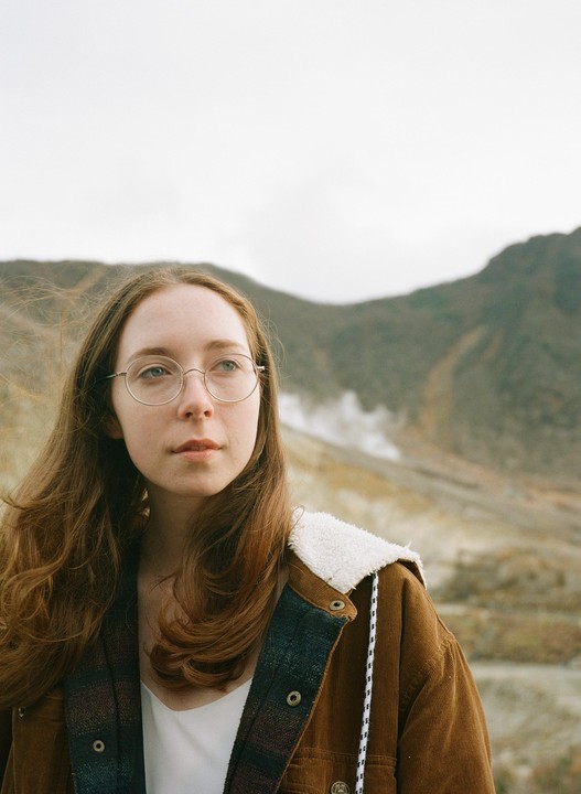 A woman with long hair and glasses wears a brown jacket with a white sherpa collar and looks to the right, with a mountain and steam in the background.