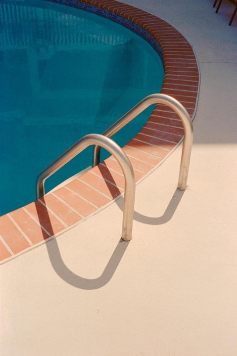 A metal ladder with shadows on the ground leads into a swimming pool with blue water and a brick edge.