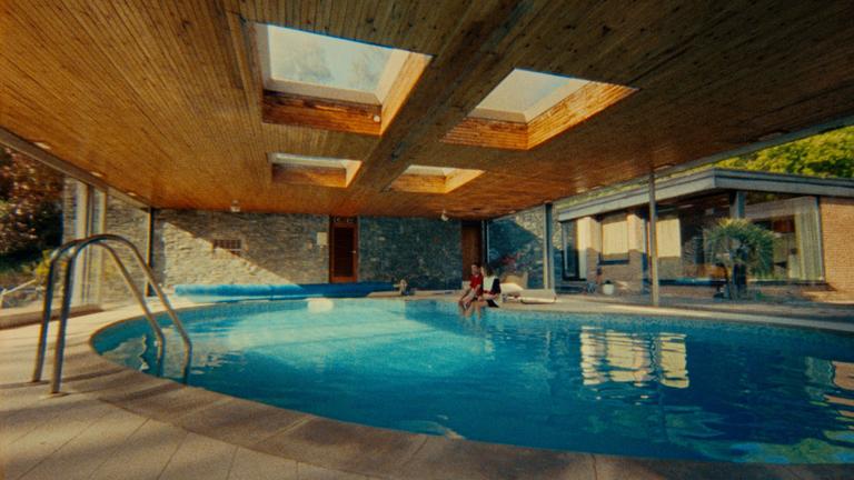 A couple sits by an indoor swimming pool with a wooden ceiling and skylights.
