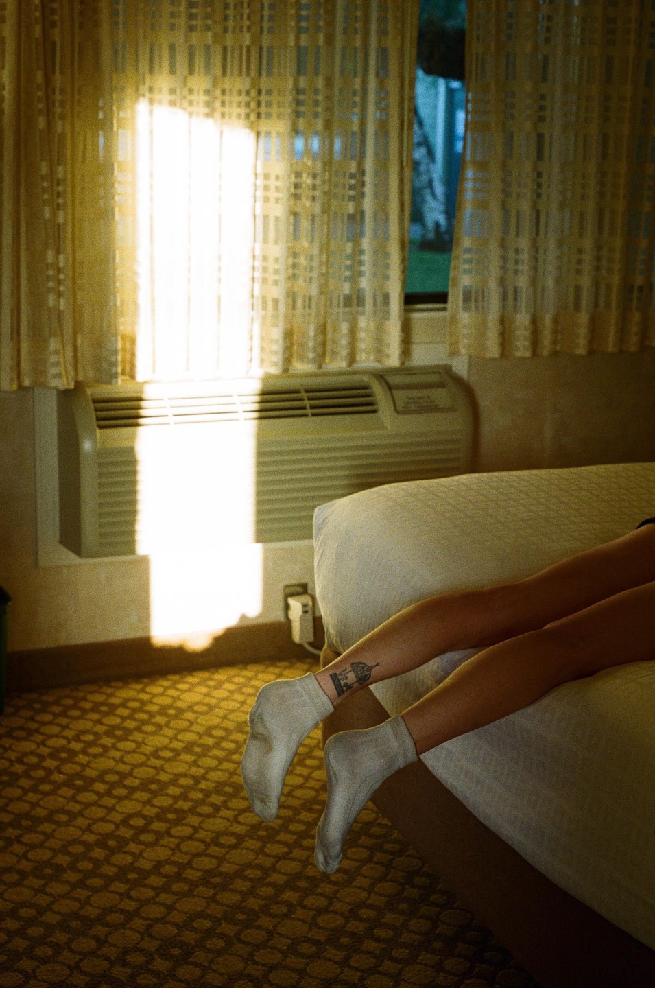 A person's legs and feet in white socks are visible next to a bed, with sunlight streaming through a window with patterned curtains.
