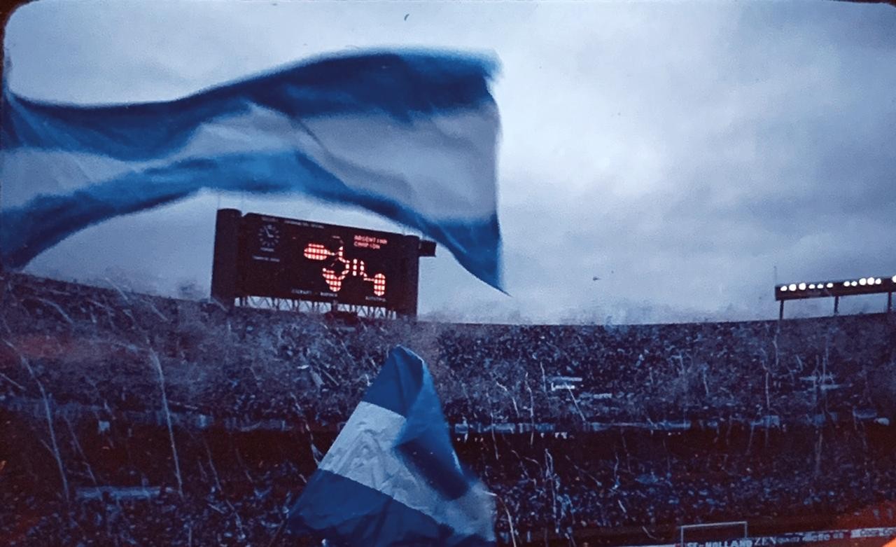 Two blue and white flags wave over a stadium filled with spectators and confetti, with a scoreboard visible in the background.