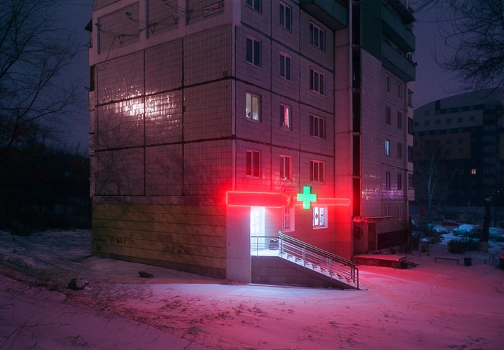 A building with a red neon sign and a green cross is illuminated at night, with snow covering the ground.