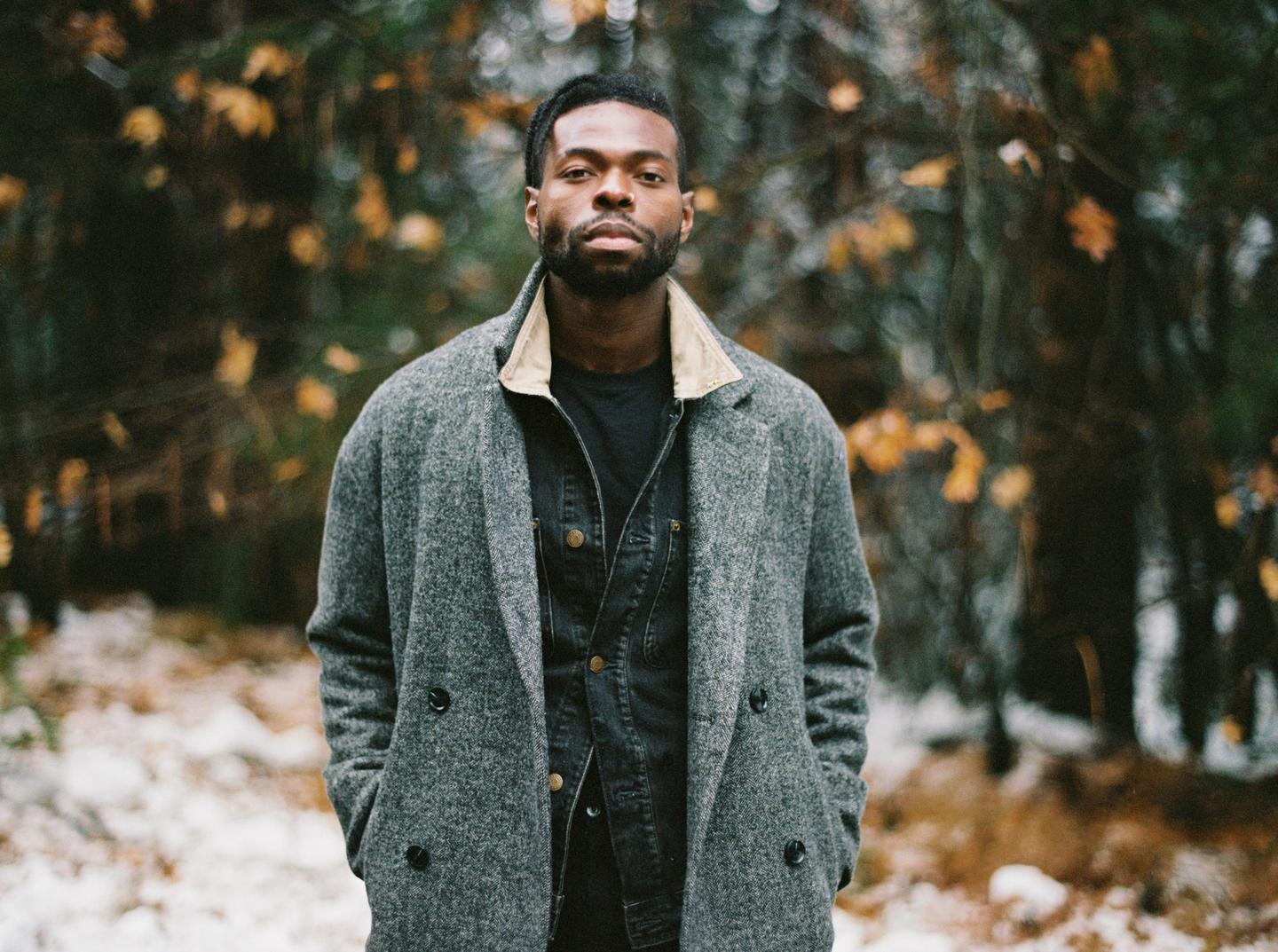 A man wearing a gray coat and a denim jacket stands in a forest with patches of snow on the ground.