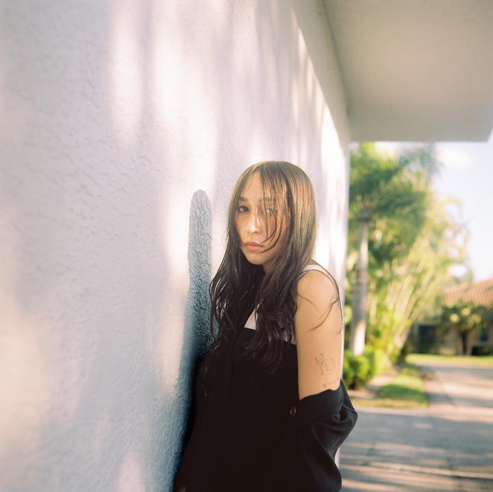 A woman with long hair stands against a white wall with shadows from palm trees.