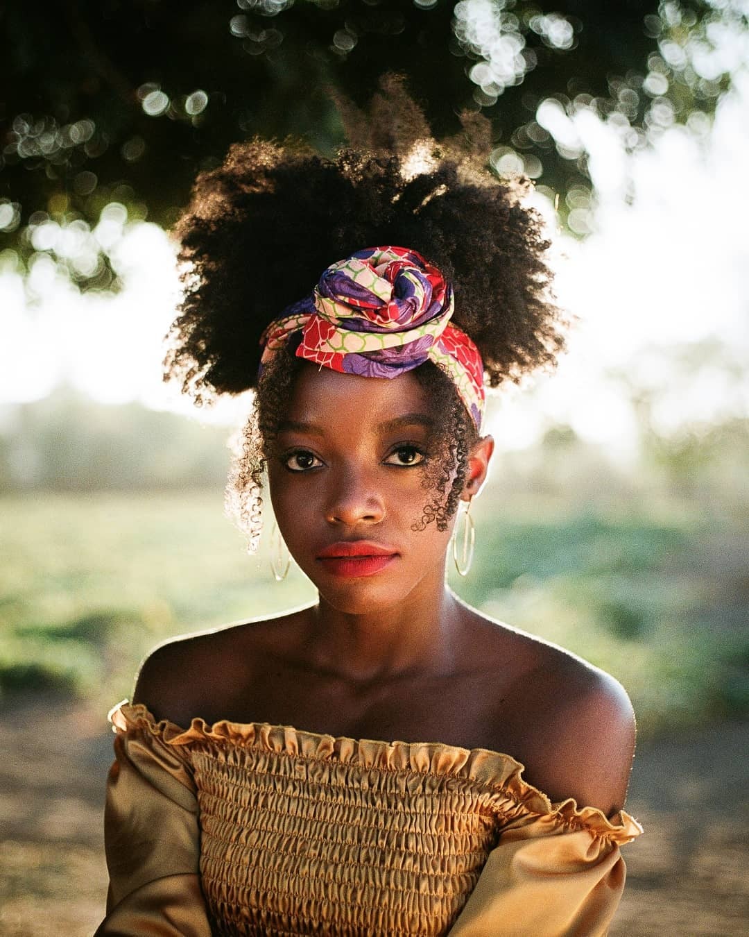 A woman with a colorful head wrap and a gold top looks directly at the viewer.