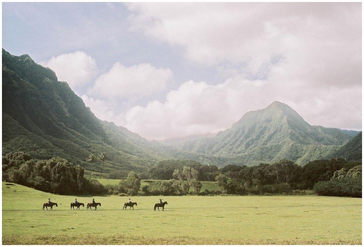 Five people on horseback ride across a green field with mountains and trees in the background.