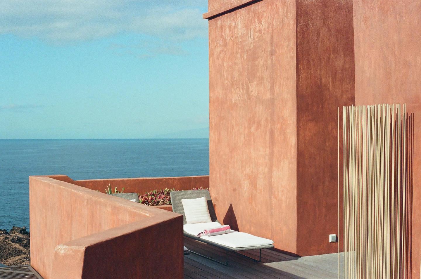 A lounge chair with a towel and pillow sits on a wooden deck next to a terracotta wall overlooking the ocean.