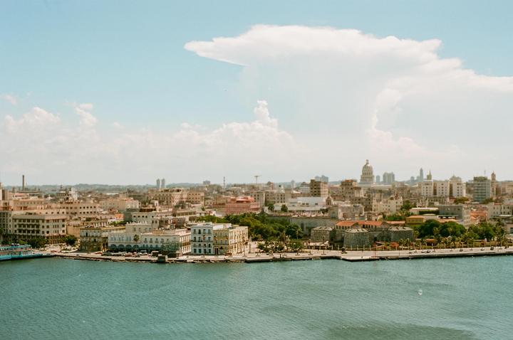 A wide shot shows the Havana cityscape with a large body of water in the foreground and a cloudy sky above.