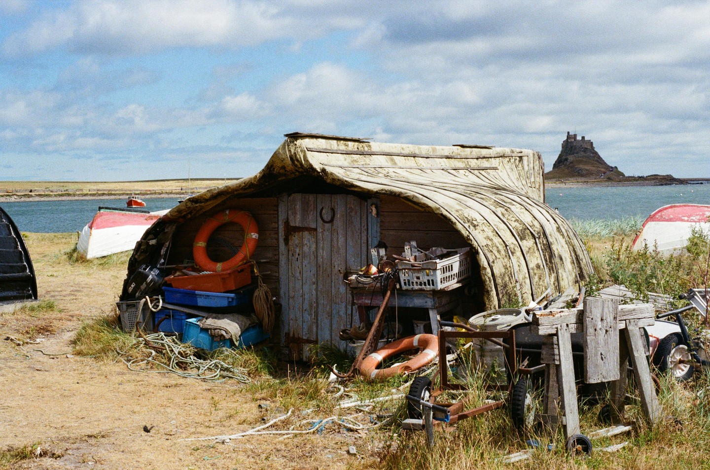 A boat shed made from an overturned boat sits on a grassy shore with a castle on an island in the background.