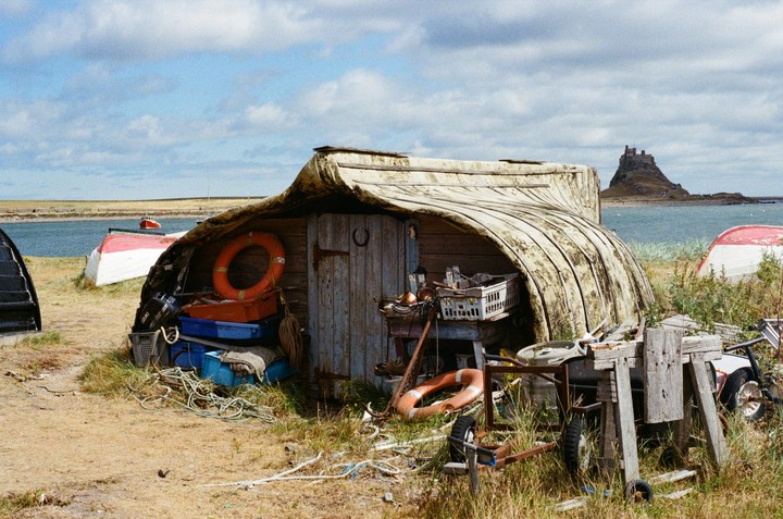 A boat shed made from an overturned boat sits on a grassy shore with a castle on an island in the background.
