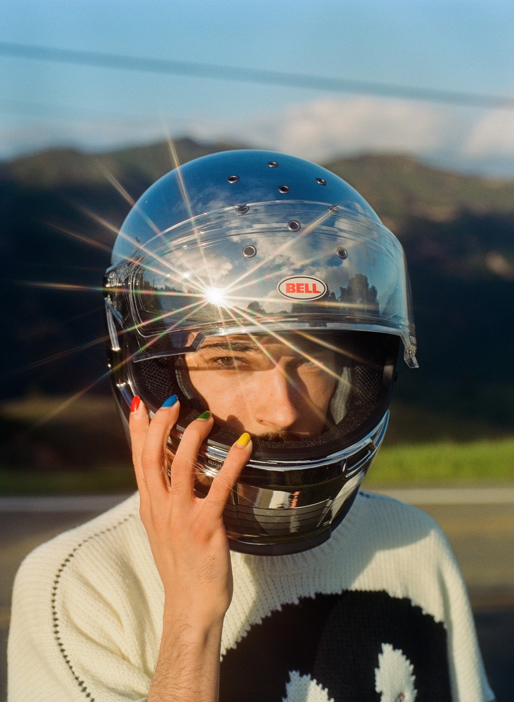 A man wearing a shiny motorcycle helmet with a sun flare on the visor touches his face with a hand with painted fingernails.