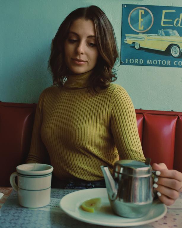 A woman in a yellow turtleneck sits at a diner table with a cup and a teapot with a lemon slice on a plate.